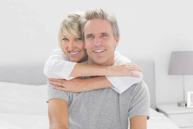 Two adults smiling, with one embracing the other from behind.